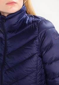 Tmavě modrá péřová bunda s vysokým límcem, prošívaným vzorem ve tvaru šipky a kruhovým logem na levém rukávu. Hladký, lehký materiál.