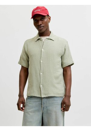 Homme portant une chemise à manches courtes boutonnée texturée vert clair, un jean bleu clair et une casquette rouge avec un texte blanc, debout devant un fond uni.