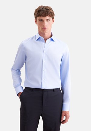 Homme portant une chemise bleue claire à manches longues boutonnée et un pantalon foncé, debout avec une main dans la poche sur fond blanc.