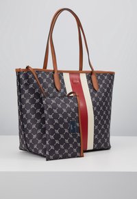 Námořnická taška přes rameno s potiskem, zdobená hnědými koženými detaily a červeným a bílým středovým pruhem. Součástí je i malá kapsa ve stejném stylu.