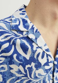 Camisa de patrón floral en azul y blanco con cuello. La tela es texturizada y ligera, con grandes motivos abstractos.