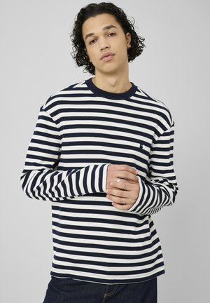 Jeune personne aux cheveux bouclés portant un t-shirt à manches longues rayé bleu marine et blanc, debout, les mains jointes, devant un fond gris uni.