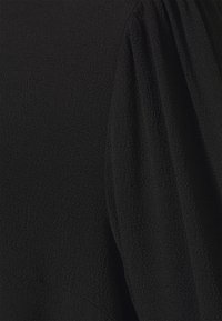 Tissu noir texturé avec des ondulations subtiles, présentant un détail de manche bouffante à l'épaule, créant une silhouette détendue.