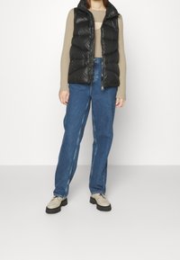 Gilet piumino nero con colletto alto, chiusura a zip, indossato sopra un top beige a coste. Jeans dritti azzurri chiari e scarpe beige con suole nere.