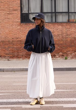 Žena oblečená v námornícky modrej bunde na zips, bielej dlhej sukni, zlatých teniskách a námornícky modrej šiltovke stojaca na ulici pred tehlovou stenou.