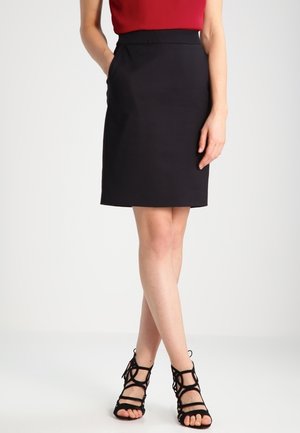 Femme portant une jupe noire jusqu'aux genoux, un haut rouge et des sandales à talons hauts noires à brides, debout avec une main dans la poche et les jambes croisées.