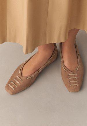 Pieds portant des ballerines pointues en daim beige avec des détails en boucle argentée, partiellement couverts par une jupe midi beige, debout sur un sol clair.