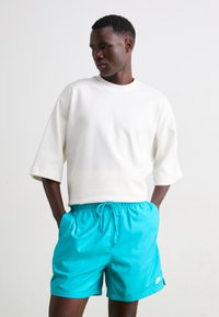 Camisa blanca de manga corta combinada con pantalones cortos turquesa brillante que cuentan con una cintura elástica y cordón, mostrando un pequeño detalle de logo.