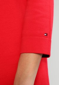 Pull rouge avec une manche côtelée et un logo discret en noir, blanc et rouge. Texture douce et design ajusté.