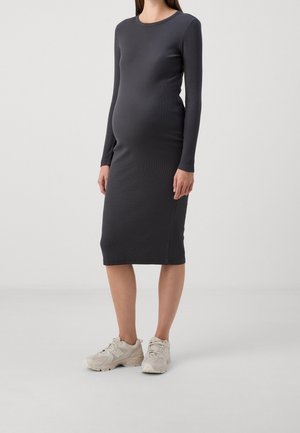 Gravid kvinde iført en tætsiddende, sort midi-kjole med lange ærmer og beige sneakers, stående på en ensfarvet lys grå baggrund.
