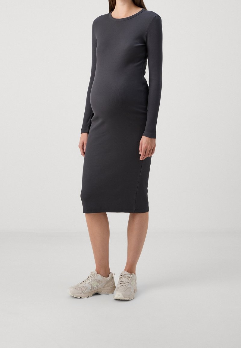 Femme enceinte portant une robe midi noire ajustée à manches longues et des baskets beiges, debout sur un fond gris clair uni.