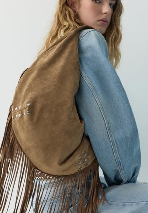 Jeune personne aux longs cheveux blonds portant une veste en jean claire et un grand sac à dos marron en daim avec des franges et des clous métalliques.