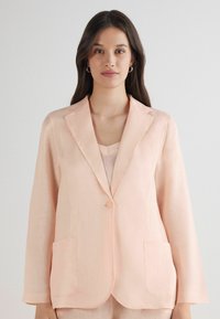 Světle růžový lněný blazer s klopovými límci, zapínáním na jeden knoflík a dvěma předními kapsami. Hladká textura a volný střih.