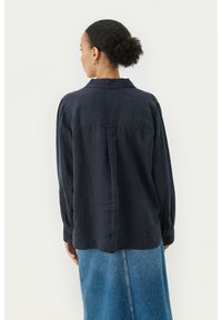 Navy blue, long-sleeve button-up shirt with a collar and back yoke detail. Worn with a knee-length denim skirt. Smooth texture.