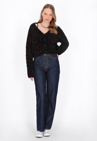 Cardigan nero texturizzato con macchie multicolori, scollatura a V e bottoni, abbinato a jeans aderenti blu scuro a vita alta e sneakers bianche.