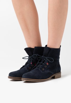 Bottes à lacets en daim bleu marine montant jusqu'à la cheville, avec des semelles marron, portées sur des jambes nues, sur un fond blanc uni.
