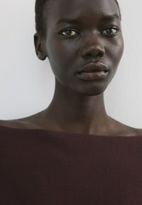 Primer plano de una persona de piel oscura y cabello corto que lleva una blusa oscura sin hombros, con un fondo claro y liso.