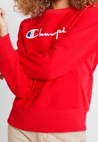 Rode sweatshirt van katoen met lange mouwen. Bevat geborduurd "Champion" logo in het wit, met een blauwe accent in de letter "C."