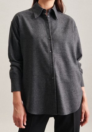 Femme portant une chemise à manches longues ample de couleur gris foncé avec col et boutons, ainsi qu'un pantalon noir, debout devant un fond neutre.