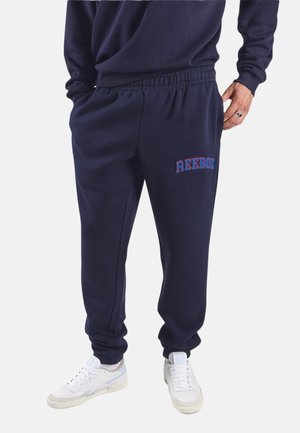 Man draagt marineblauwe Reebok joggingbroek met elastische boorden en witte sneakers, handen in de zakken, staand tegen een witte achtergrond.
