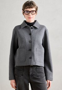 Casaco cinza curto feito de um tecido macio, com gola, seis botões pretos e dois bolsos frontais. Combinado com jeans escuros.