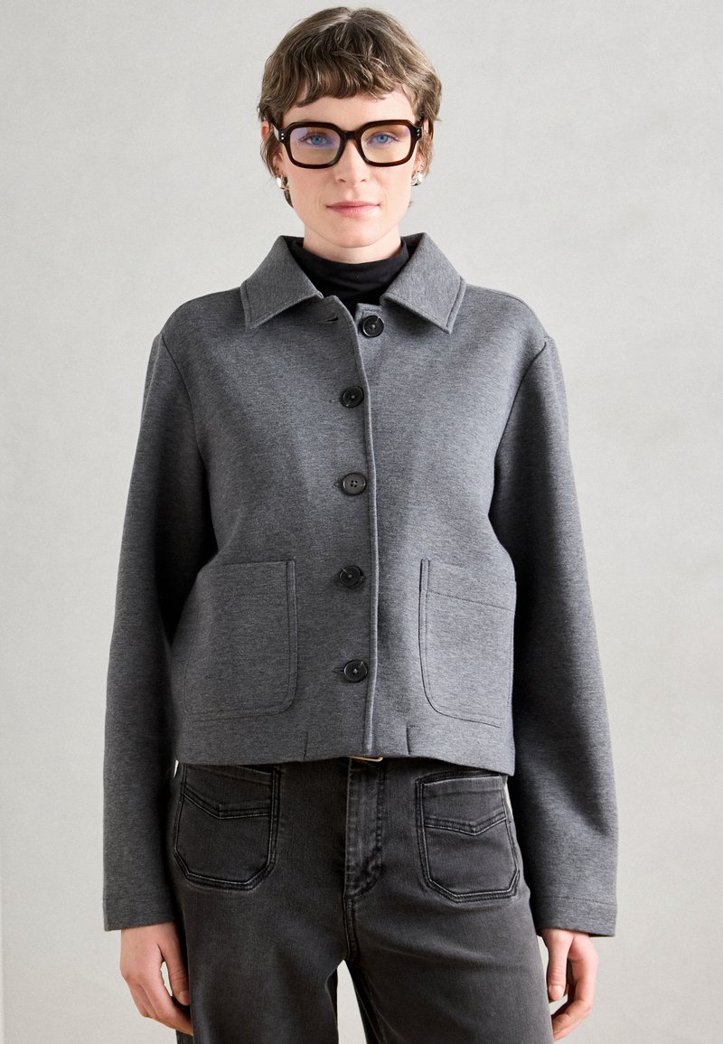 Casaco cinza curto feito de um tecido macio, com gola, seis botões pretos e dois bolsos frontais. Combinado com jeans escuros.