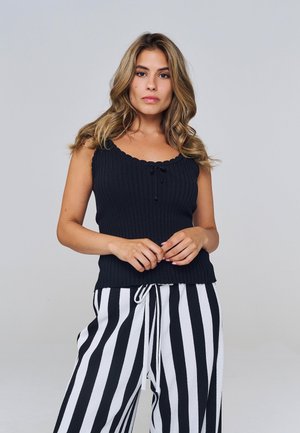 Naine laineliste blondide juustega, kandes musta varrukateta soonikutoppi ja kõrge vöökohaga must-valgete vertikaalsete triibuliste püksteid, seistes neutraalsel taustal.