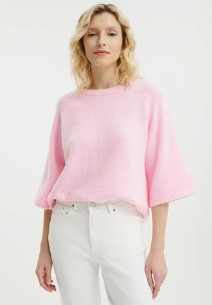 Helles rosa Strickpullover mit kurzen, weiten Ärmeln und rundem Halsausschnitt, kombiniert mit weißen, hoch taillierten Skinny-Jeans. Weicher, strukturiertes Material.