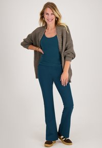 Overmaatse, gestructureerde taupe cardigan over een teal tanktop, gecombineerd met bijpassende teal broek en bruine schoenen met crèmekleurige strepen.