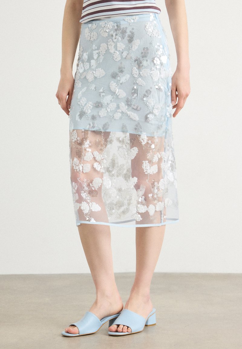 Jupe midi bleu clair avec broderie florale en sequins, tissu transparent et ourlet qui tombe sous les genoux. Associée à des mules ouvertes bleu clair.