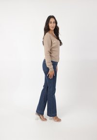 Beige textuur cardigan met knopen, gecombineerd met donkerblauwe flared jeans en transparante blokhak sandalen. Model poseert zijwaarts.