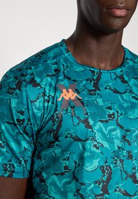 Homme portant un t-shirt de fitness à motif abstrait teal et noir avec un logo Kappa orange sur la poitrine, visible des épaules à la bouche.
