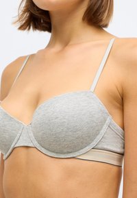 Sutiã cinza acolchoado com alças finas ajustáveis, apresentando uma textura suave e uma faixa elástica beige em contraste ao longo da parte inferior do busto.