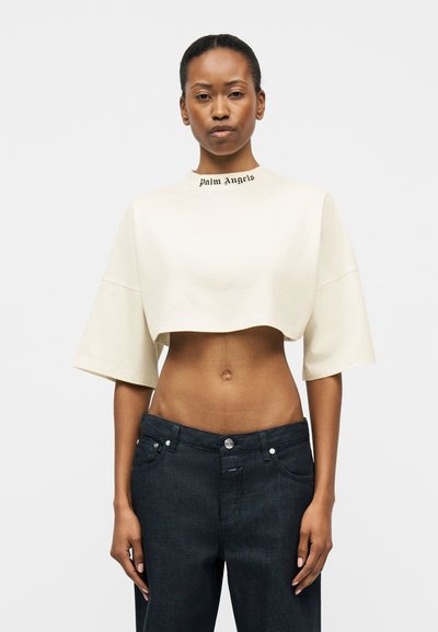 Γυναίκα με λευκό crop top με το κείμενο "Palm Angels" στη φάσα και ψηλόμεσα σκούρα τζιν, στέκεται μπροστά σε απλό φόντο.