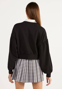Giovane donna con lunghi capelli castani indossa un maglione nero corto e una gonna plissettata bianca e nera a quadri, vista da dietro.