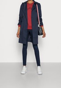 Abrigo tipo trench azul marino con botones negros, combinado con un sudadera roja, jeans oscuros y zapatillas blancas. Incluye un pequeño bolso negro con una correa de rayas.