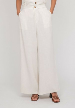 Femme portant un pantalon écru taille haute à jambes larges avec des boutons et des sandales marron à talons, les mains dans les poches, sur un fond neutre.