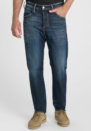 Man die donkerblauwe spijkerbroek en beige casual schoenen draagt, staand met één hand in de zak tegen een effen achtergrond.
