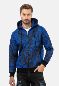 Blaues Zip-Hoodie mit schwarzen Löwen- und Tribal-Mustern, mit Kordelzugkapuze und gerippten Bündchen, aus weichem Stoff gefertigt.