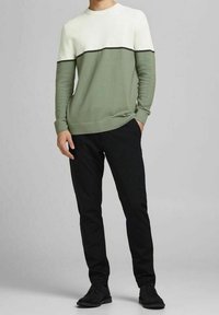 Man in een wit en olijfgroen geribd sweater, met zwarte broek en zwarte schoenen, staande met één hand in zijn zak tegen een grijze achtergrond.