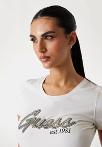 Balta medvilninė marškinė su sidabriniais rhinestones „Guess“ logotipu, apvaliu iškirpimu ir trumpomis rankovėmis. Lygus tekstūra, prigludusi forma.