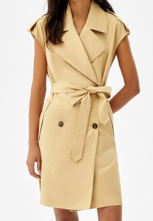 Beżowa sukienka trencz bez rękawów z szerokimi klapami, zapinana na dwa rzędy guzików, z paskiem wiązanym w talii, noszona przez osobę o długich ciemnych włosach.