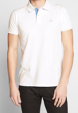 Mann trägt ein weißes Poloshirt mit einem blauen inneren Kragen und kleinem GANT-Logo auf der Brust, kombiniert mit schwarzen Hosen.