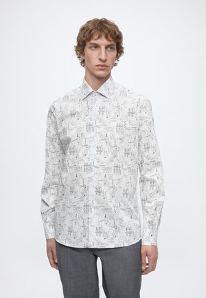 Jeune homme portant une chemise blanche boutonnée avec des motifs croquis noirs et un pantalon gris, debout devant un fond blanc uni.