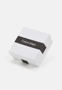 Boîte-cadeau en carton blanc avec un bandeau noir arborant le logo "Calvin Klein". Vue de dessus en angle, montrant un design minimaliste.