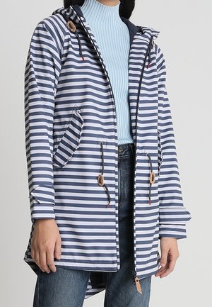 Femme portant une veste à capuche à rayures horizontales bleu marine et blanc par-dessus un col roulé côtelé bleu clair et un jean bleu.