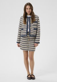 Femme portant un ensemble veste et jupe en tweed beige et bleu marine avec des boutons dorés, une chemise blanche et des chaussures plates noires, debout devant un fond uni.