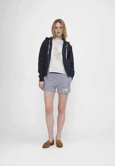 Sweatshirt azul-marinho com fecho e cordões brancos, t-shirt bege com um gráfico de cavalo e calções às riscas com um logótipo. Sapatilhas de camurça castanhas.