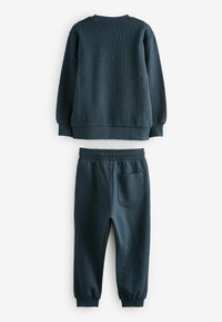 Tamsiai mėlynas džemperis ir derančios džinsinės kelnės, pagamintos iš minkštos medžiagos. Džemperis turi raukšlėtas rankoves; džinsinės kelnės turi užpakalinį kišenę.