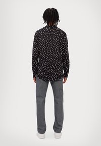 Černá košile s bílým vzorem hvězd, dlouhé rukávy, límeček na knoflíky a šedé džíny. Model stojí zády a má na sobě casual obuv.